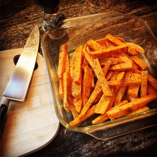 Seasoned sweet potato fries in a glass baking dish next to a knife on a cutting board.