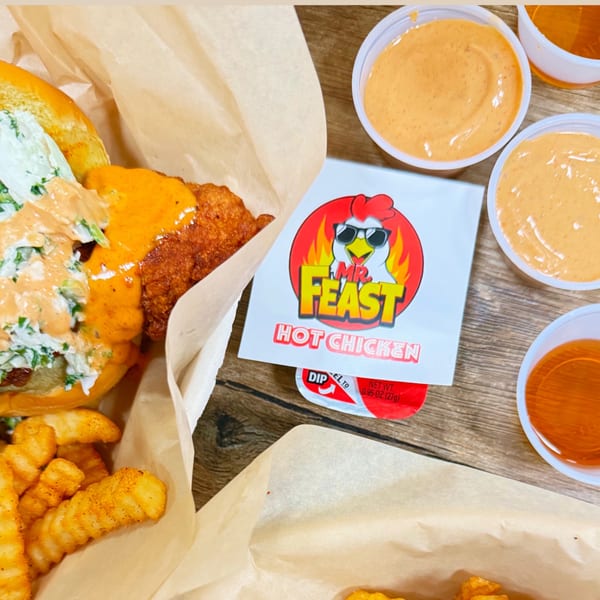 Mr. Feast Hot Chicken sandwich with fries, sauces, and branded packaging on a wooden table.