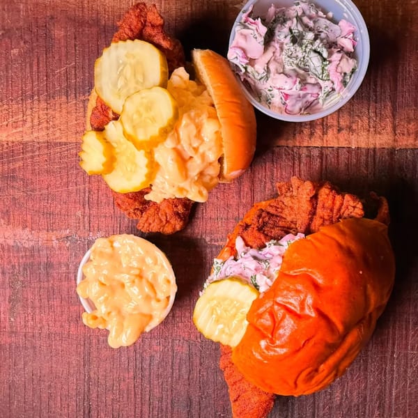 Two Mr Feast Hot Chicken sandwiches with pickles, mac and cheese, and coleslaw on a wooden surface.