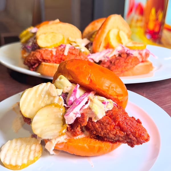 Three Mr Feast Hot Chicken sandwiches with fried chicken, coleslaw, and pickles on white plates.