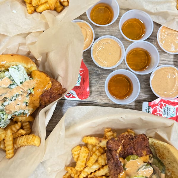 Two Mr Feast Hot Chicken sandwiches with fries, various dipping sauces, and ketchup on a wooden table.